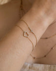 An angled view of a wrist showcasing a thin gold chain bracelet with a sparkling sideways cross paired with a heart bracelet.