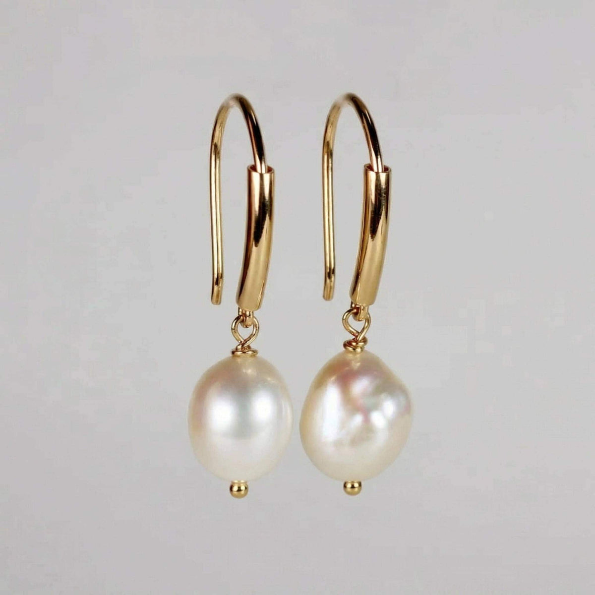 A pair of gold dangle earrings featuring leverback hooks and white baroque freshwater pearls against a plain light gray background.
