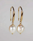 A pair of gold dangle earrings featuring leverback hooks and white baroque freshwater pearls against a plain light gray background.
