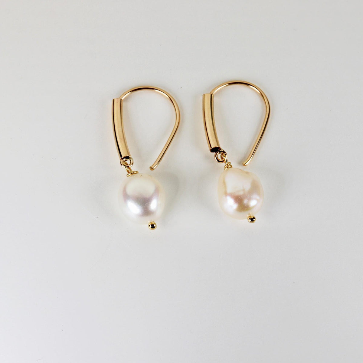 The same pair of gold-filled and white baroque pearl dangle earrings laid flat on a solid light background, viewed from an angled perspective.