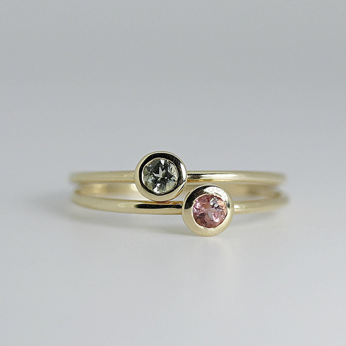 Two thin gold rings stacked vertically on a neutral surface, one holding a bezel-set light green gemstone and the other a bezel-set pink gemstone.