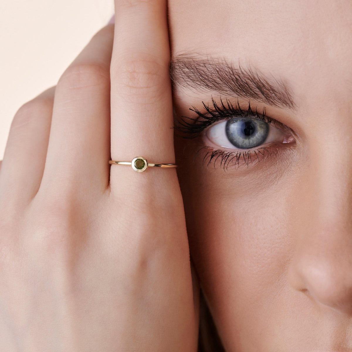 Close-up of a person holding their hand near their face, wearing a thin gold ring with a bezel-set green stone.