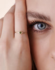 Close-up of a person holding their hand near their face, wearing a thin gold ring with a bezel-set green stone.
