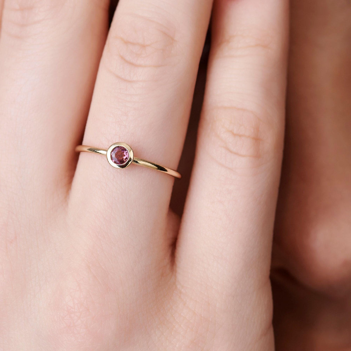 Close-up of a hand wearing a thin gold ring with a bezel-set pink gemstone.