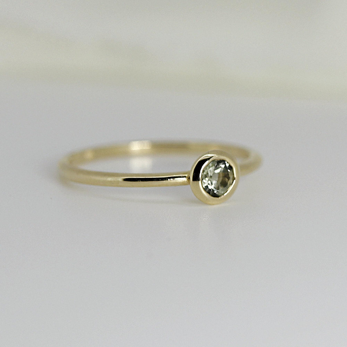 Side-angled view of a thin gold band featuring a round, bezel-set pale green gemstone.