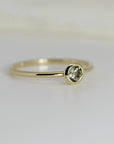 Side-angled view of a thin gold band featuring a round, bezel-set pale green gemstone.