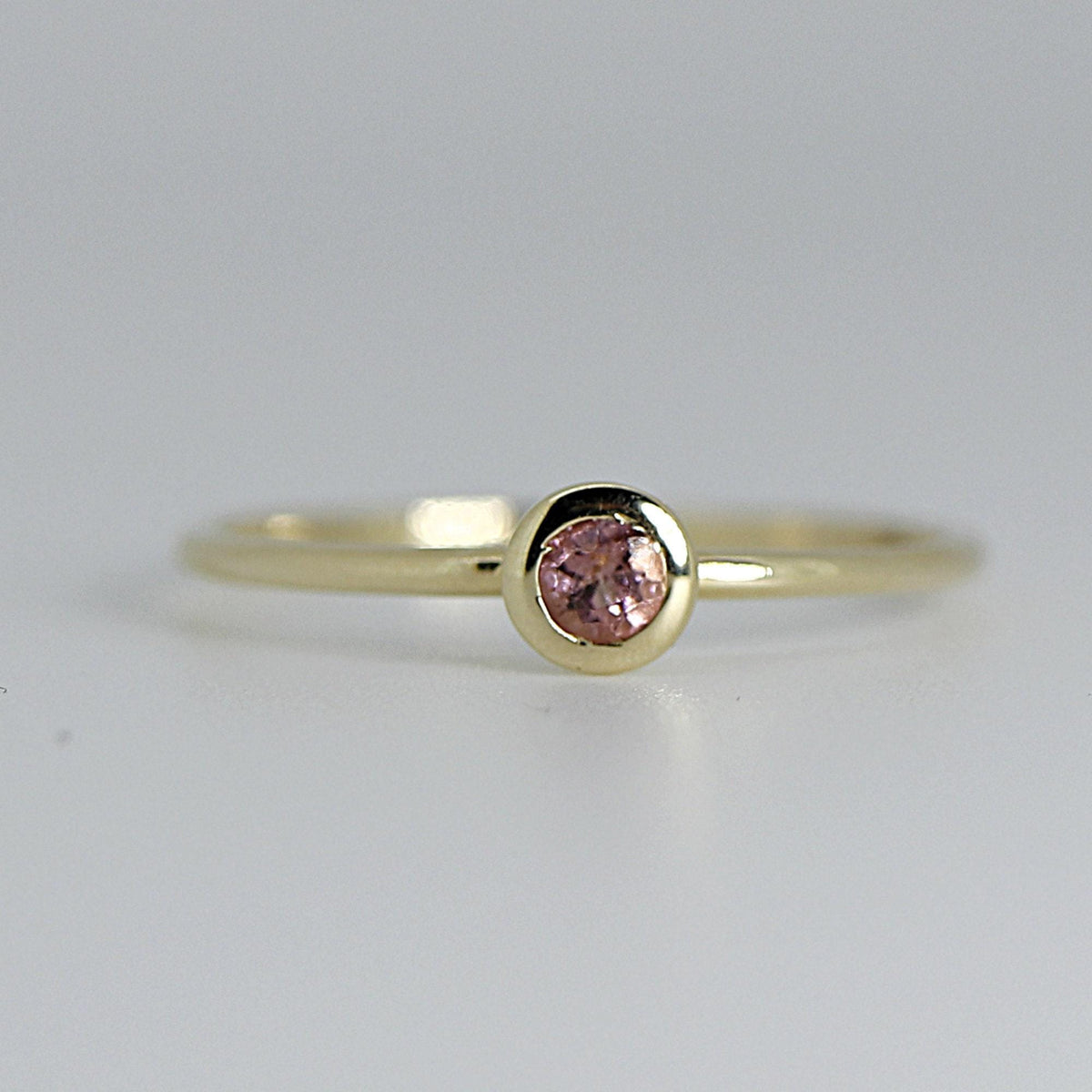 Close-up view of a thin gold ring with a round, bezel-set pink gemstone on a plain background.