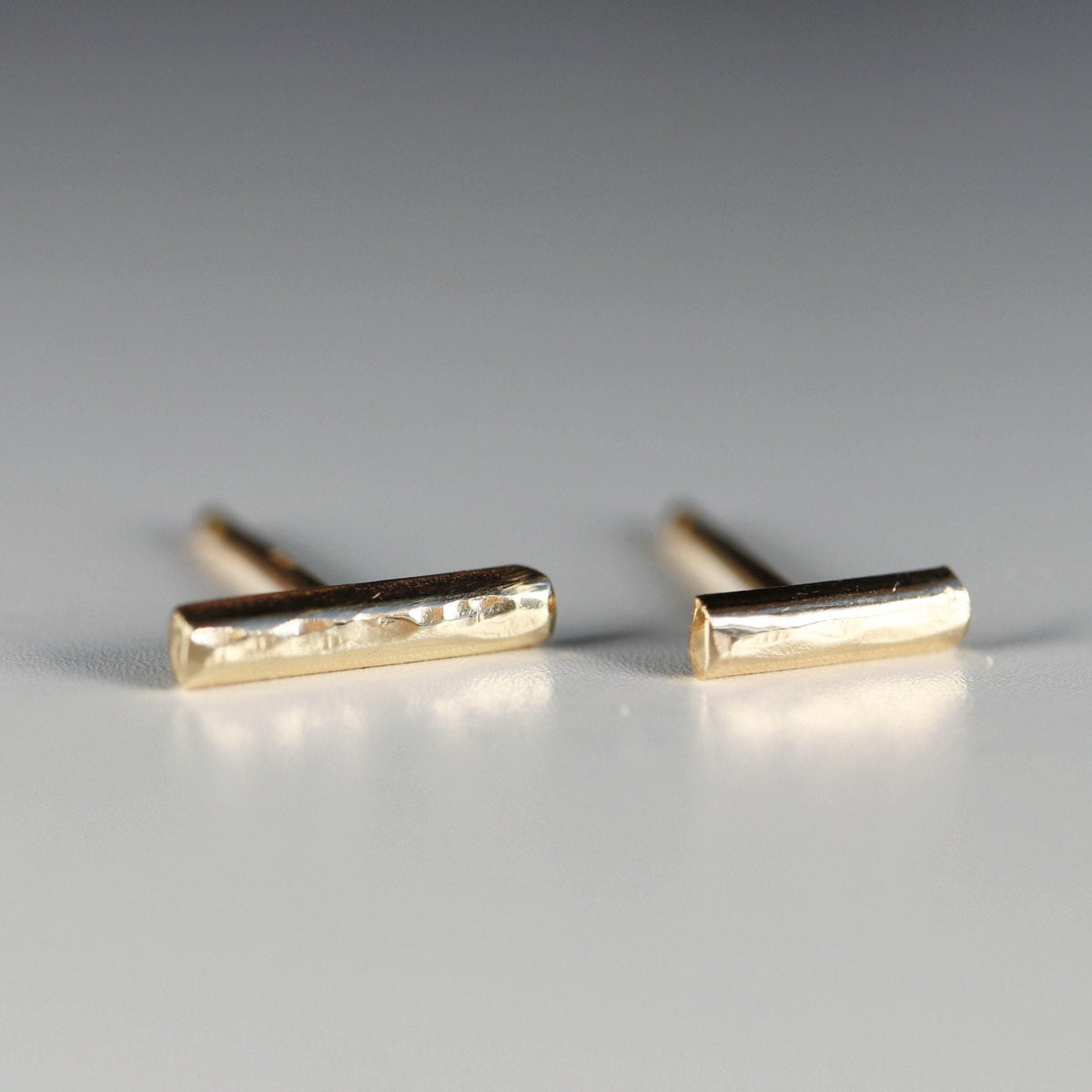 Two gold bar stud earrings with a hammered finish are arranged on a grey surface.