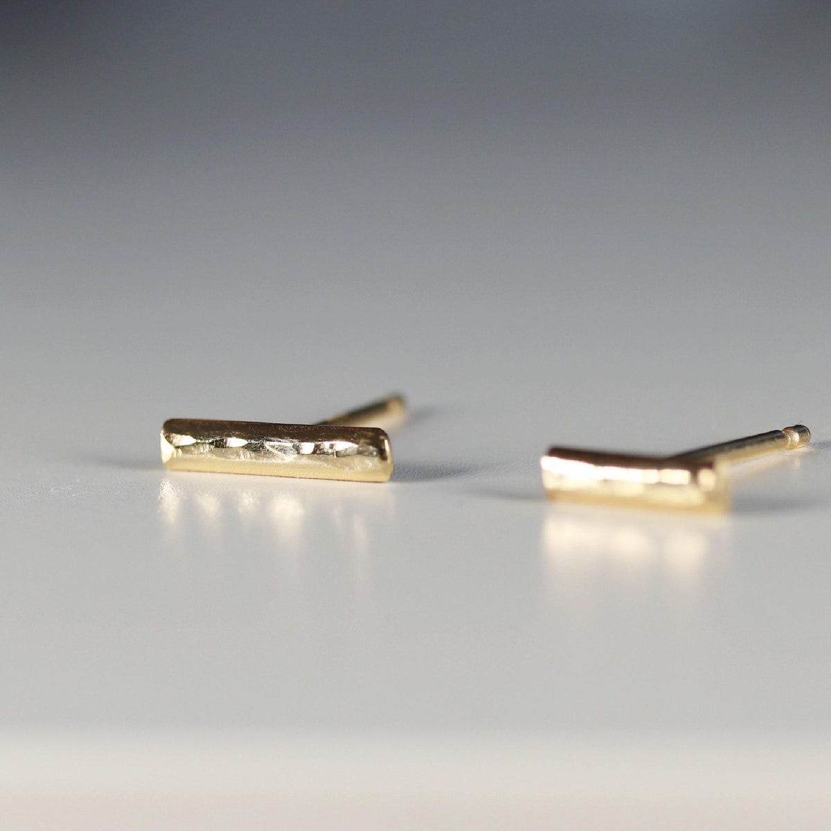 Two gold bar stud earrings are positioned on a grey surface, viewed from a low angle.