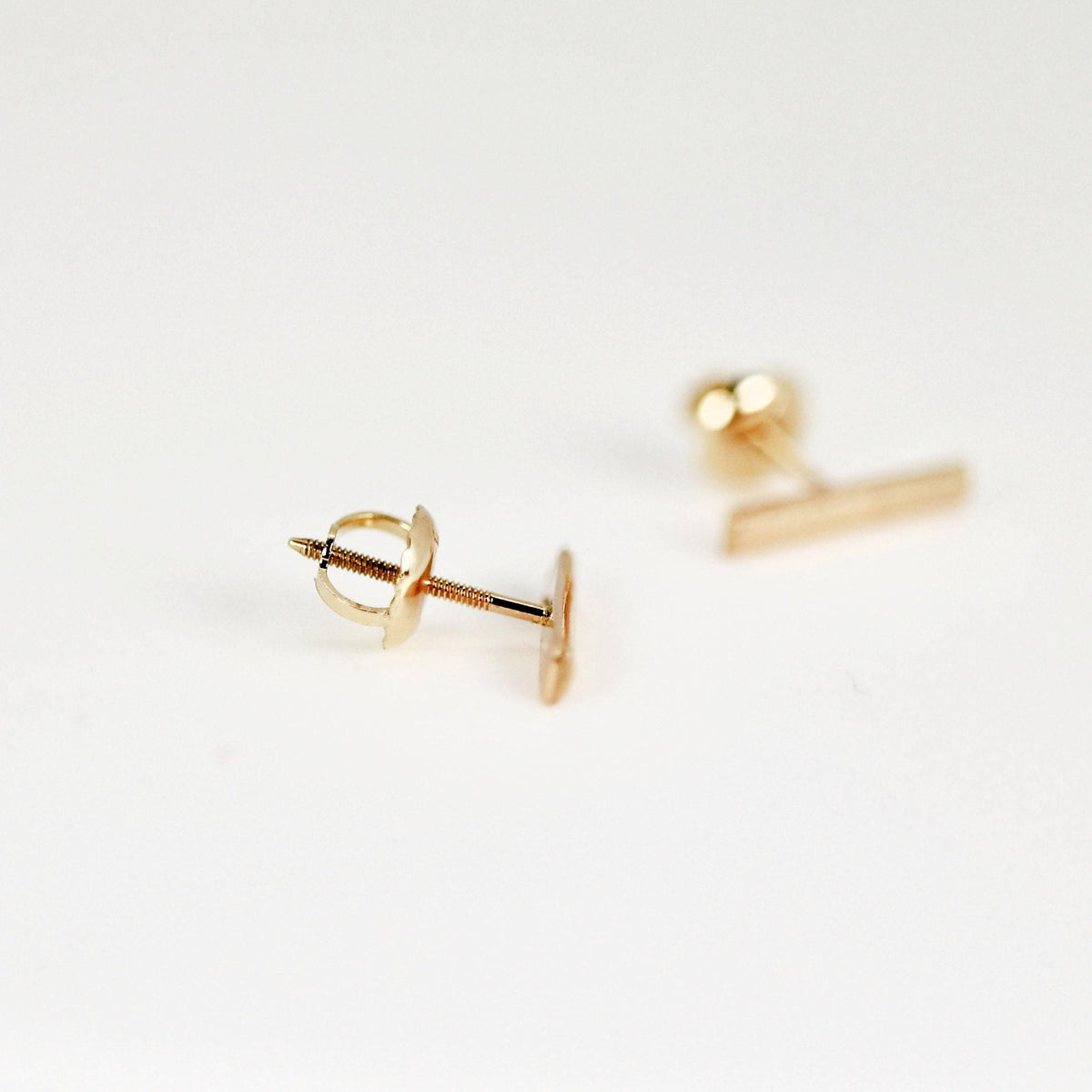 A gold bar stud earring displays its threaded post and screw back mechanism on a white surface.