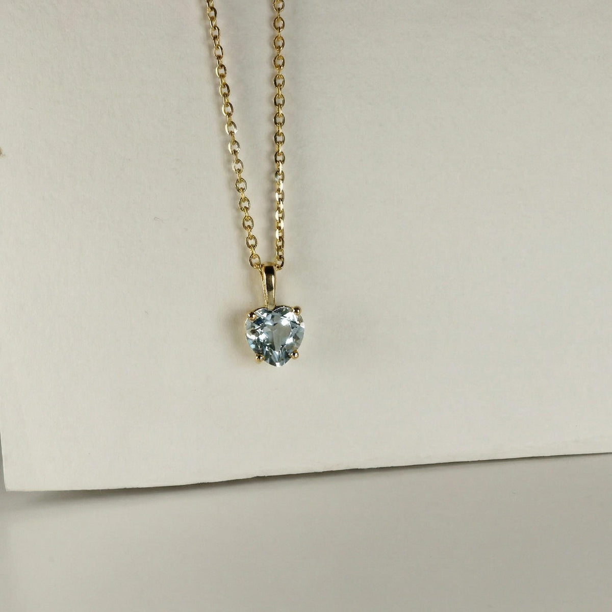 A dainty gold chain necklace with a faceted light blue heart pendant rests against a white surface.