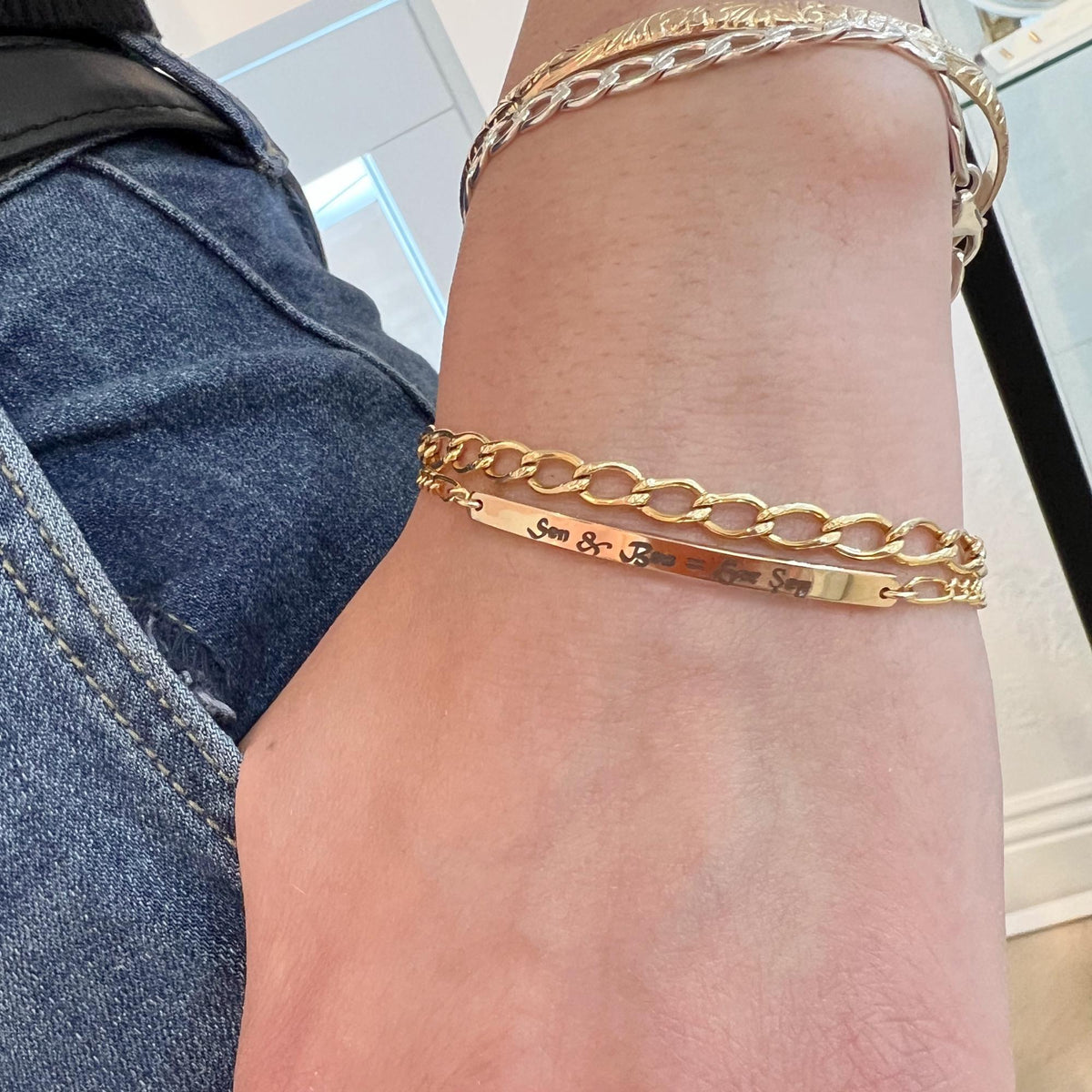 A wrist wearing an engraved gold bar bracelet with a curb chain next to other silver and gold bracelets.