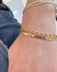 A wrist wearing an engraved gold bar bracelet with a curb chain next to other silver and gold bracelets.