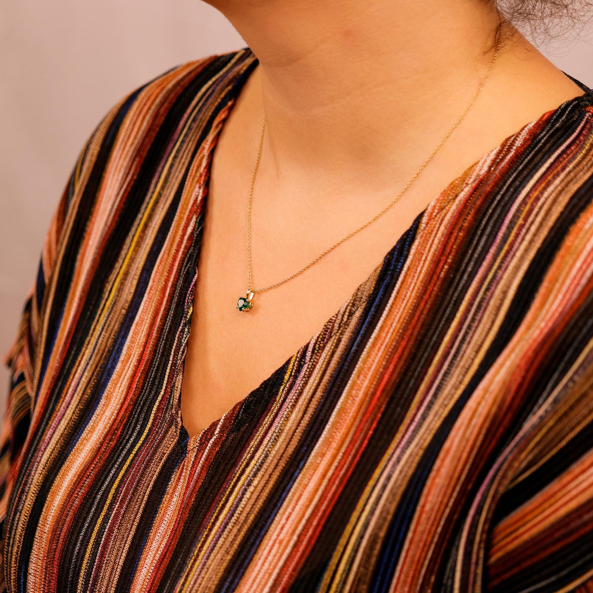 A person wearing a multicolored striped top wears a gold chain necklace with a green heart pendant.