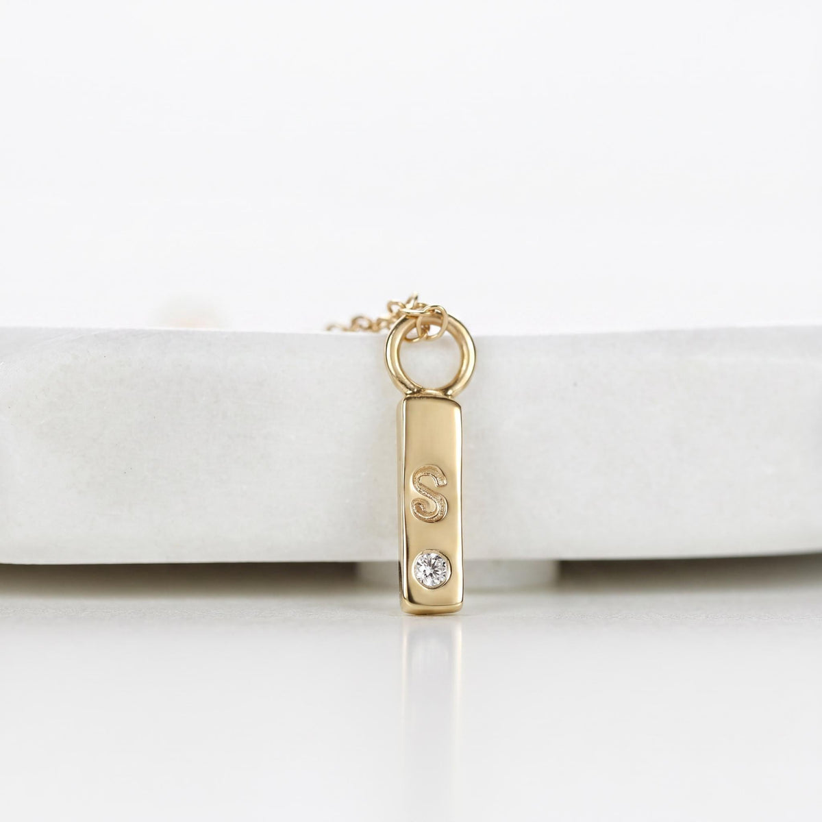 Close-up of a gold vertical bar pendant necklace resting on a white surface, featuring an engraved letter S and a small embedded diamond.