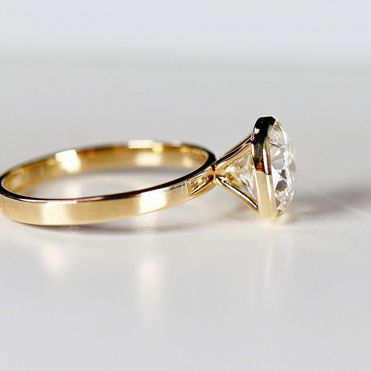 Profile view of a gold bezel-set cushion cut ring resting on a light surface, showing the gallery and band.
