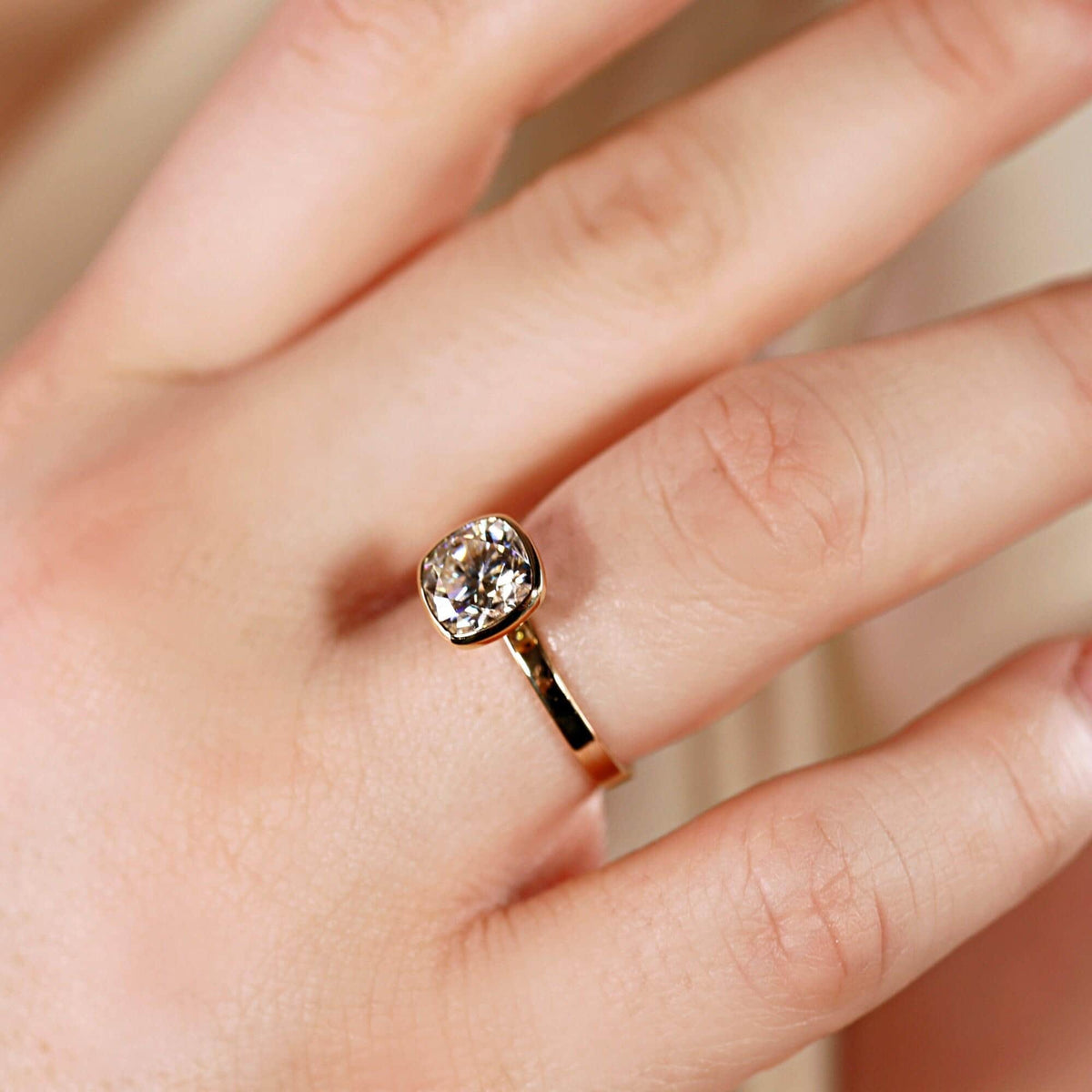 Close-up of a gold bezel-set cushion cut ring worn on a finger against a blurred background.