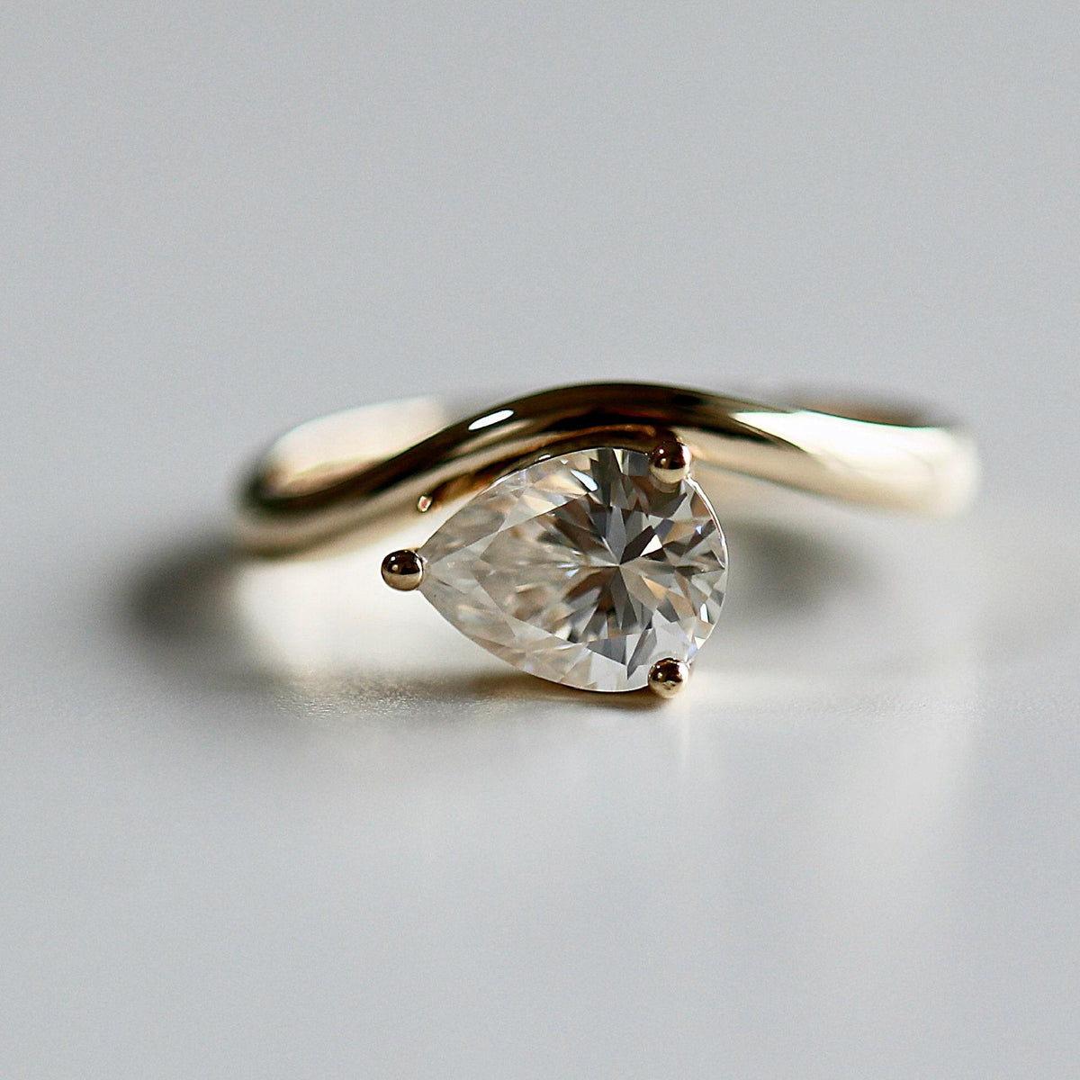 A close-up studio shot of a gold ring featuring a pear-shaped gemstone held by three prongs on a wavy band.