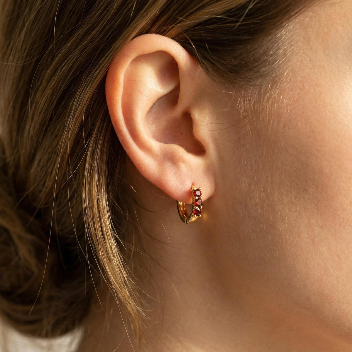 A close-up side profile of a person wearing a gold hoop earring set with three red stones in the earlobe.