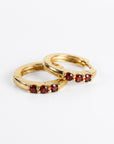 Two gold huggie hoop earrings resting on a white surface, each set with three small red round stones.