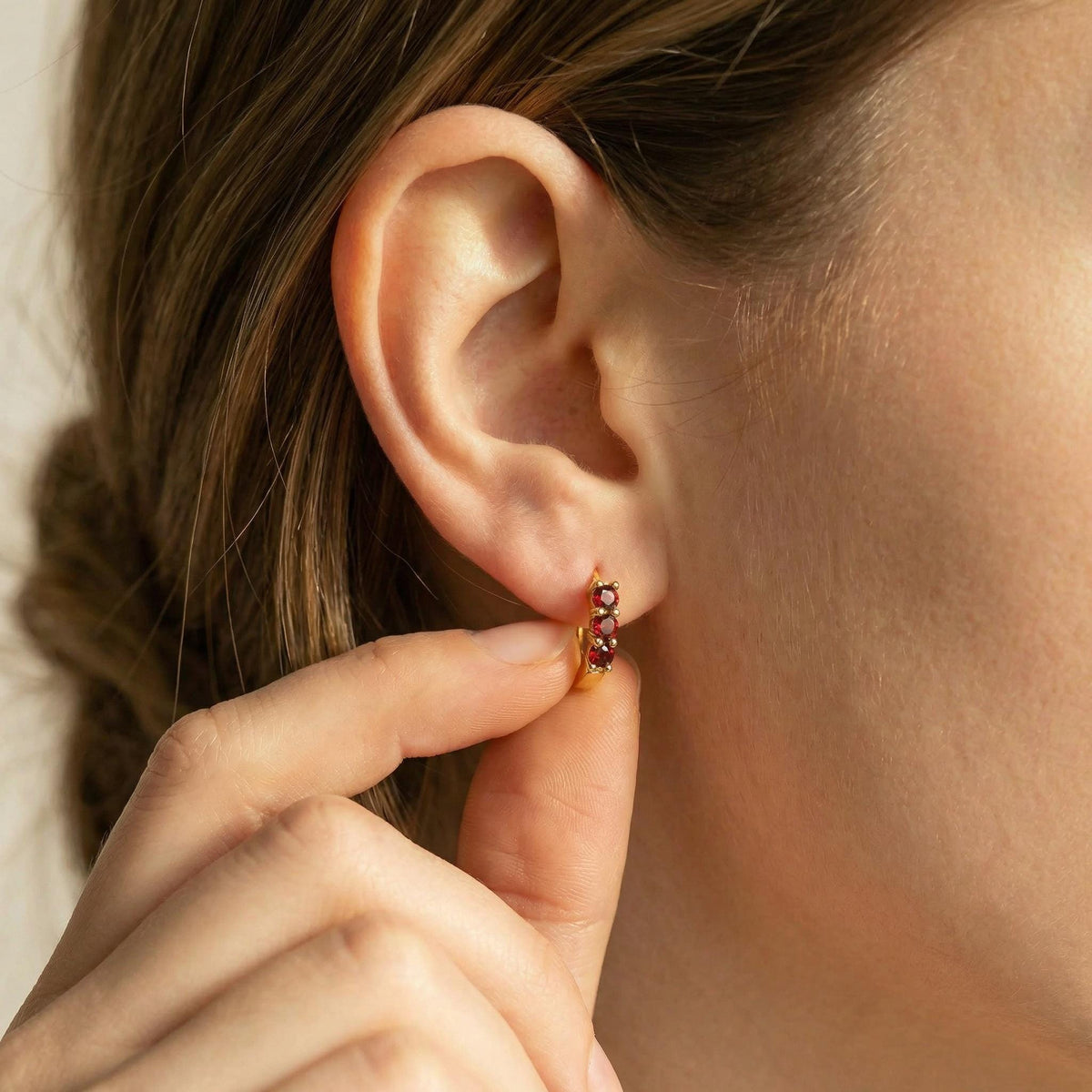 A hand lightly touches a gold huggie hoop earring worn in an earlobe, highlighting the three red stones.