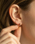 A hand lightly touches a gold huggie hoop earring worn in an earlobe, highlighting the three red stones.