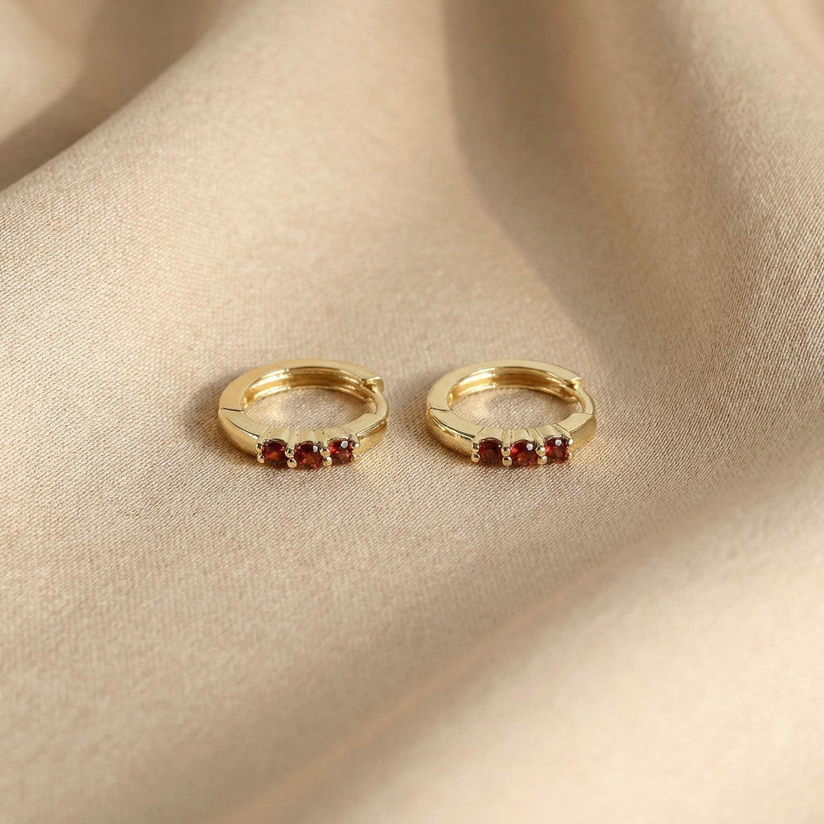 A pair of gold huggie hoop earrings with red gemstone accents resting on beige fabric.