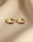 A pair of gold huggie hoop earrings with red gemstone accents resting on beige fabric.