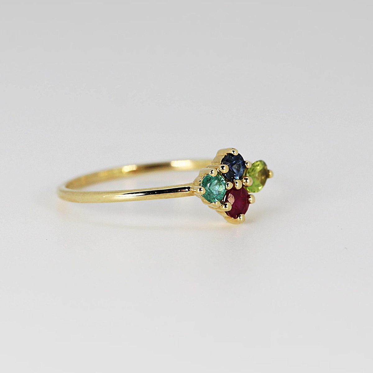 Side view of a gold cluster ring featuring red, blue, green, and light green gemstones resting on a white surface.