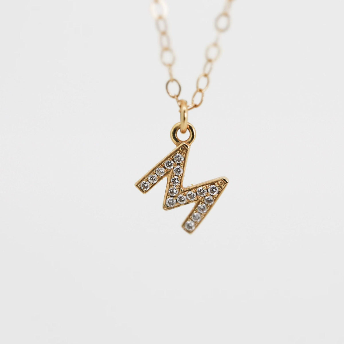 A gold chain necklace featuring a small letter M pendant embellished with diamonds, shown against a plain white background.