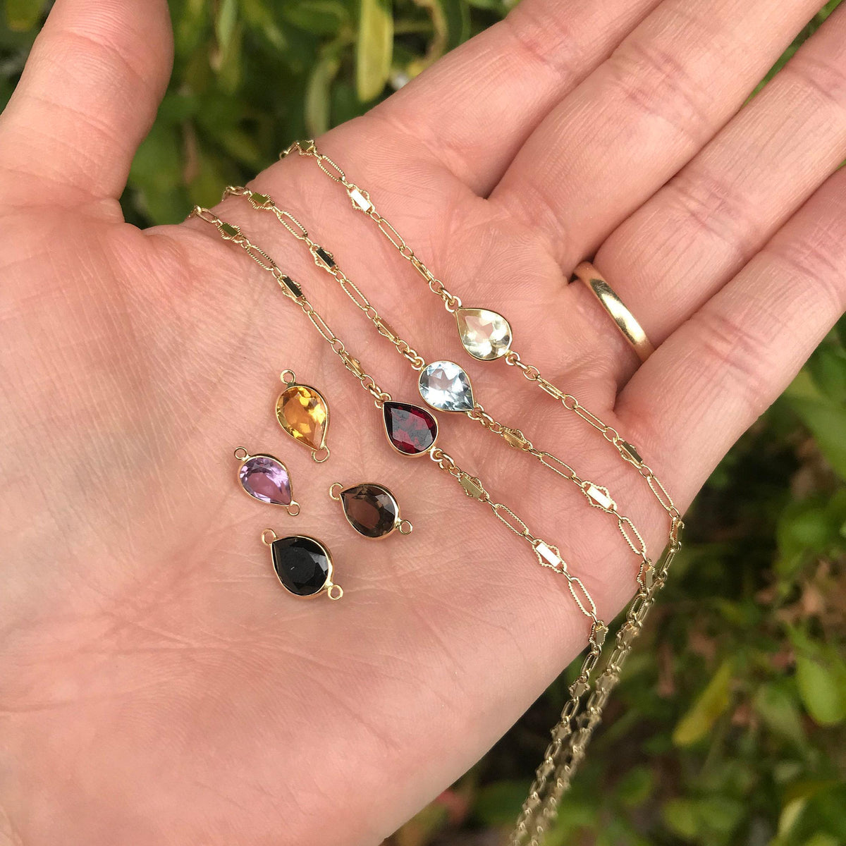 An open hand displays four gold chain necklaces with various gemstone pendants, plus five extra loose pear-shaped gemstone charms.