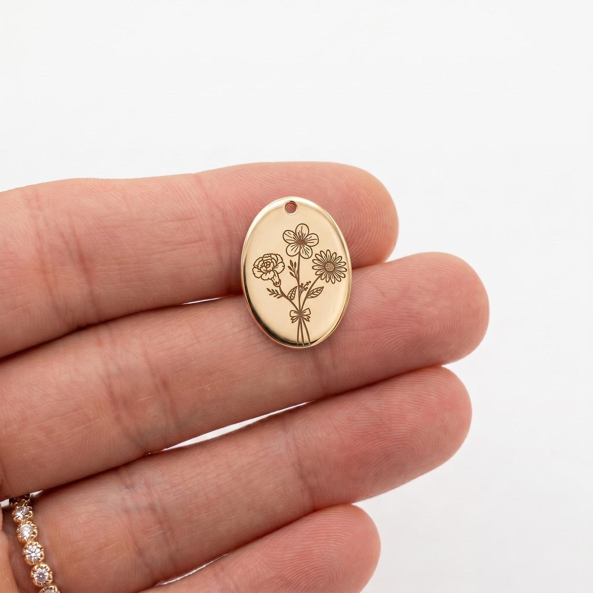 A gold-colored oval metal disc pendant held in a person's palm, featuring an engraved bouquet of three flowers tied with a small bow.