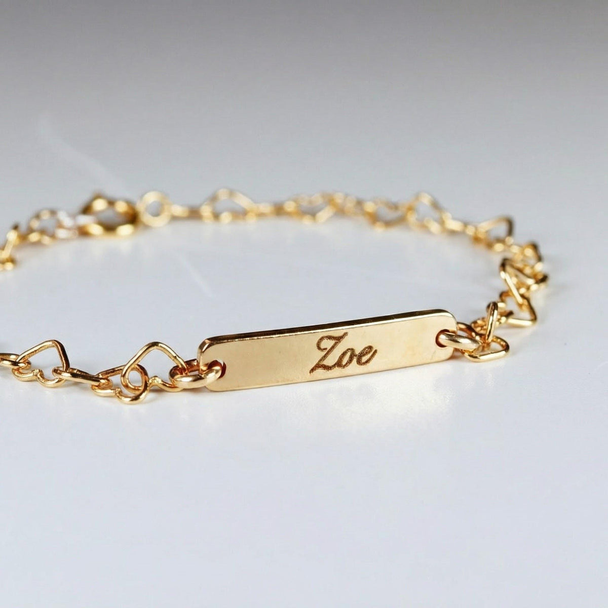 A gold bracelet featuring a rectangular bar engraved with the name Zoe and a chain made of linked hearts rests on a white surface.