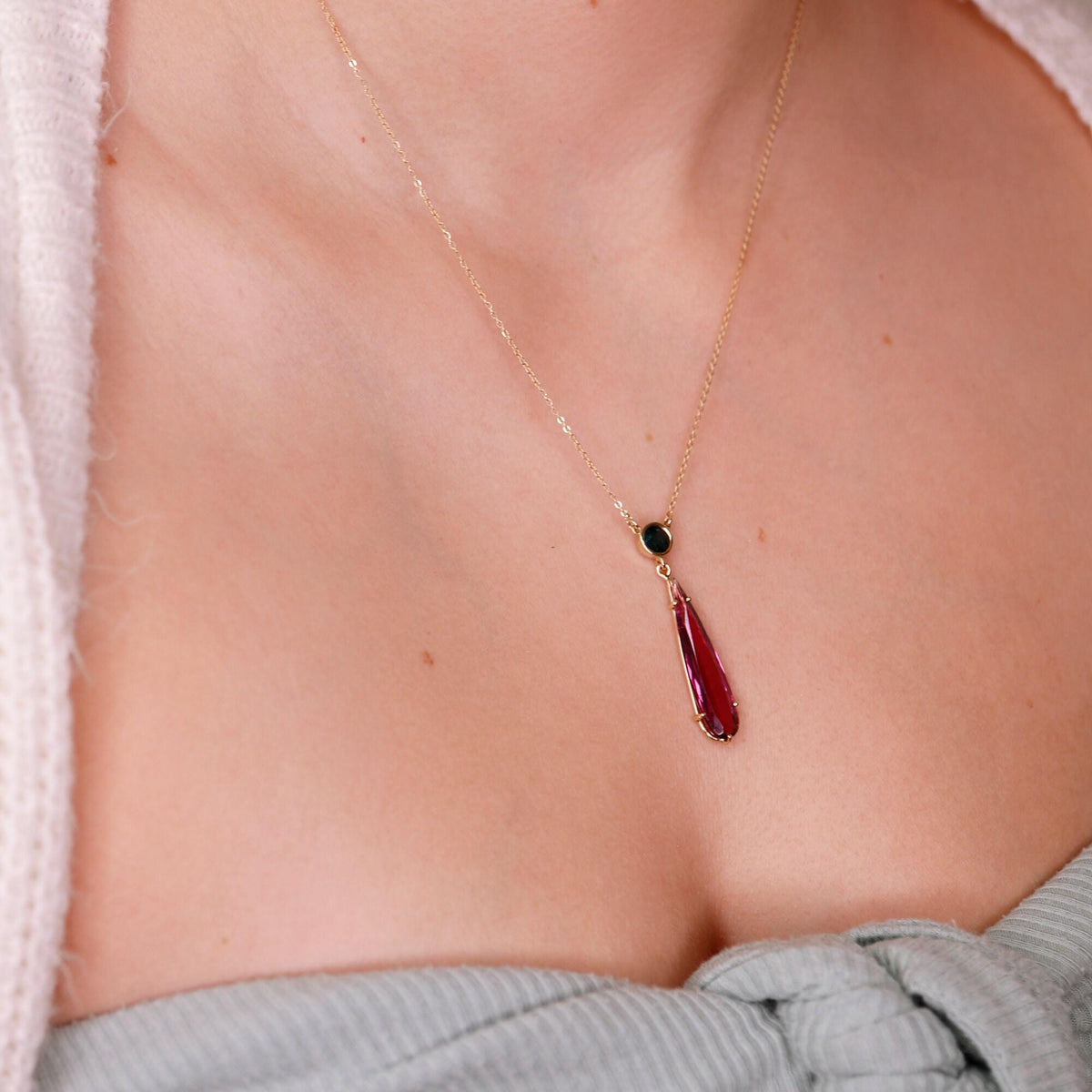A close-up of a person wearing a gold chain necklace with a dark round stone and a hanging pink teardrop gemstone pendant.