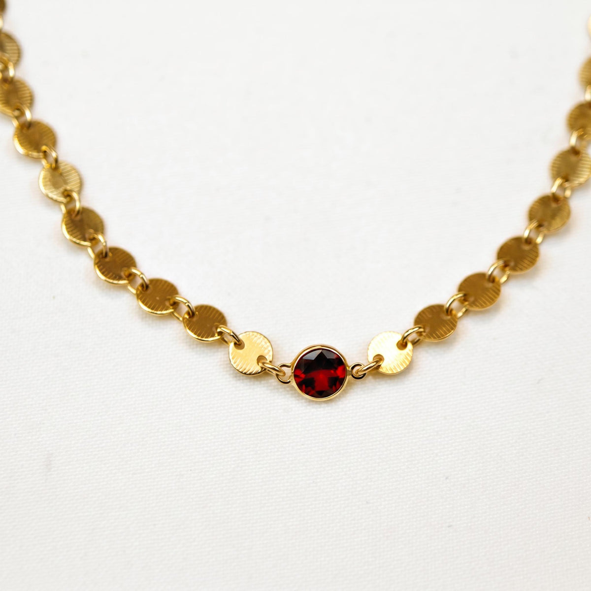 Close-up view of a gold sequin disc chain necklace with a single round dark red garnet gemstone in the center.