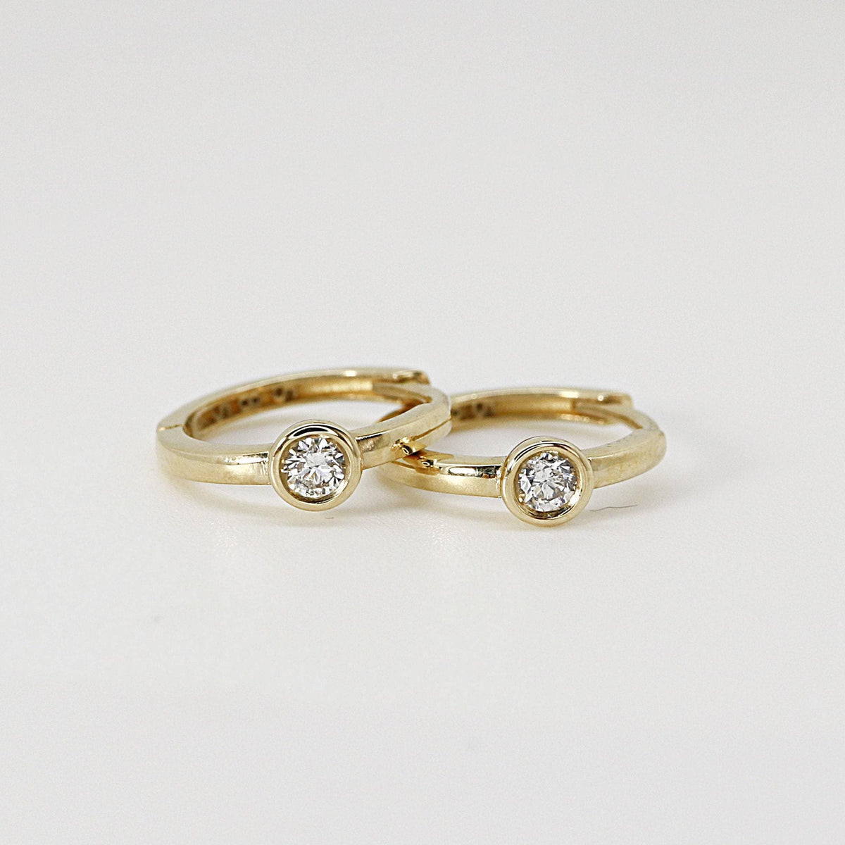 Two small gold hoop earrings with bezel-set solitaire diamonds resting side by side on a white surface.