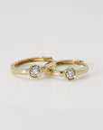 Two small gold hoop earrings with bezel-set solitaire diamonds resting side by side on a white surface.