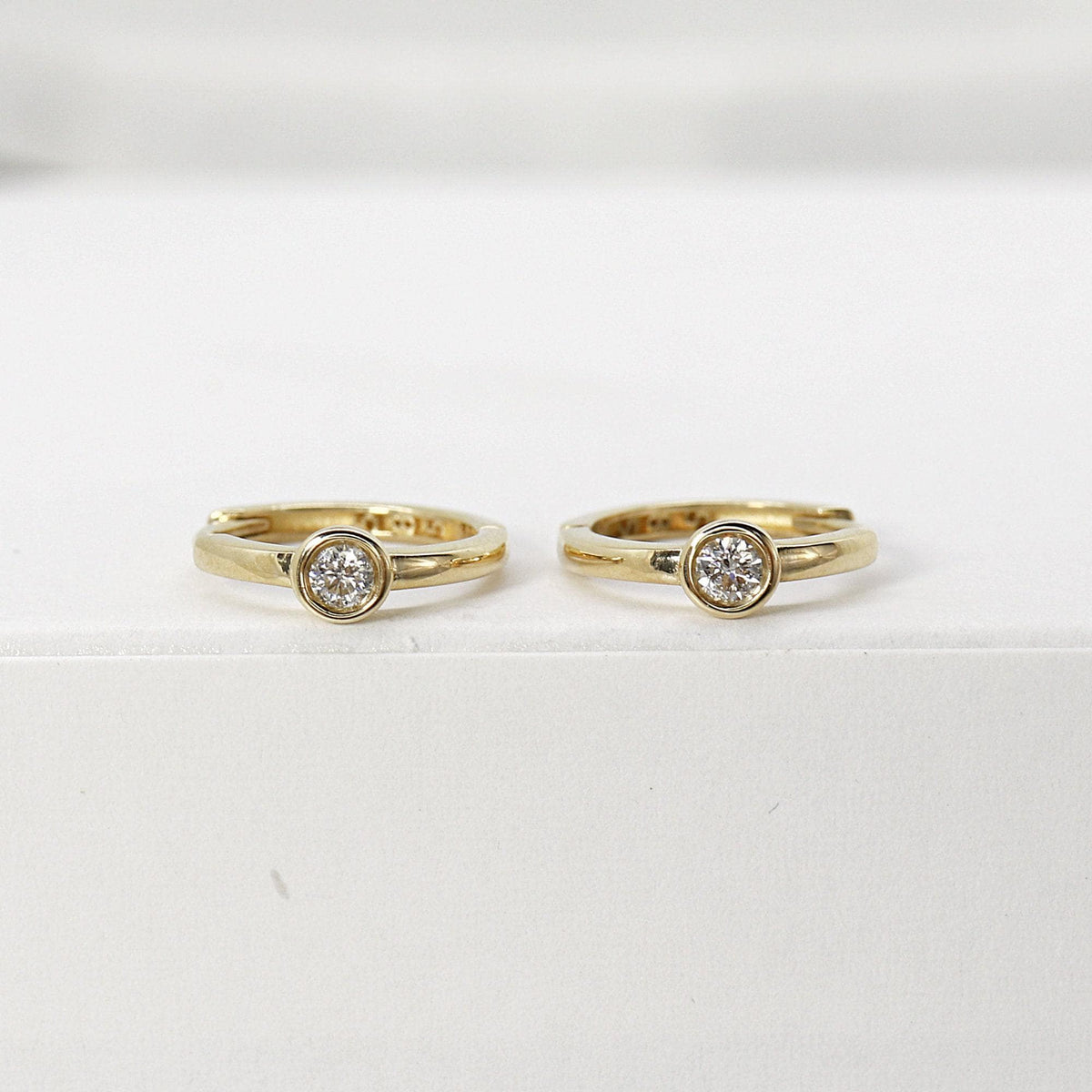 Two gold hoop earrings with bezel-set diamonds displayed upright on a white platform.