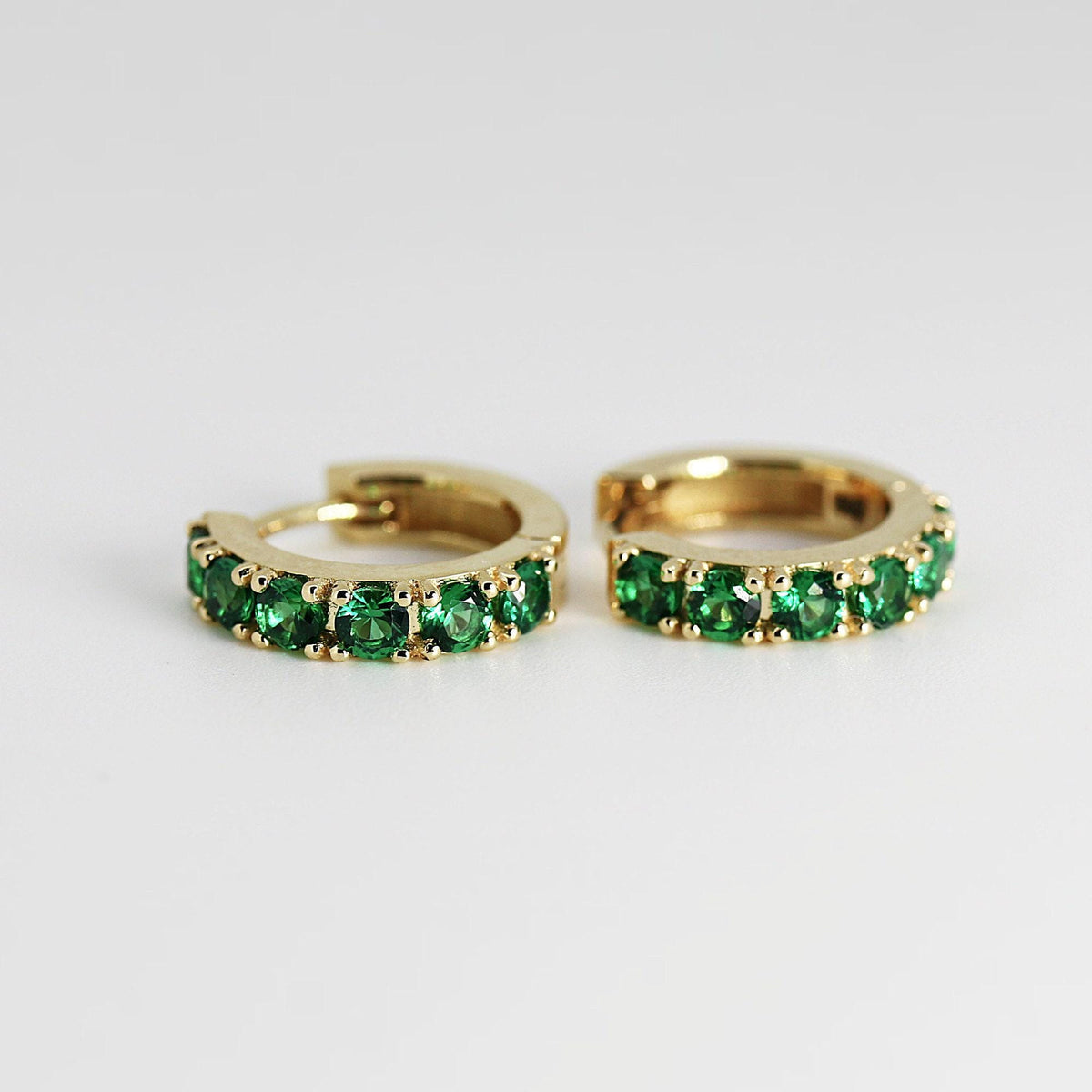 Two small gold huggie hoop earrings featuring a row of round green stones against a neutral background.