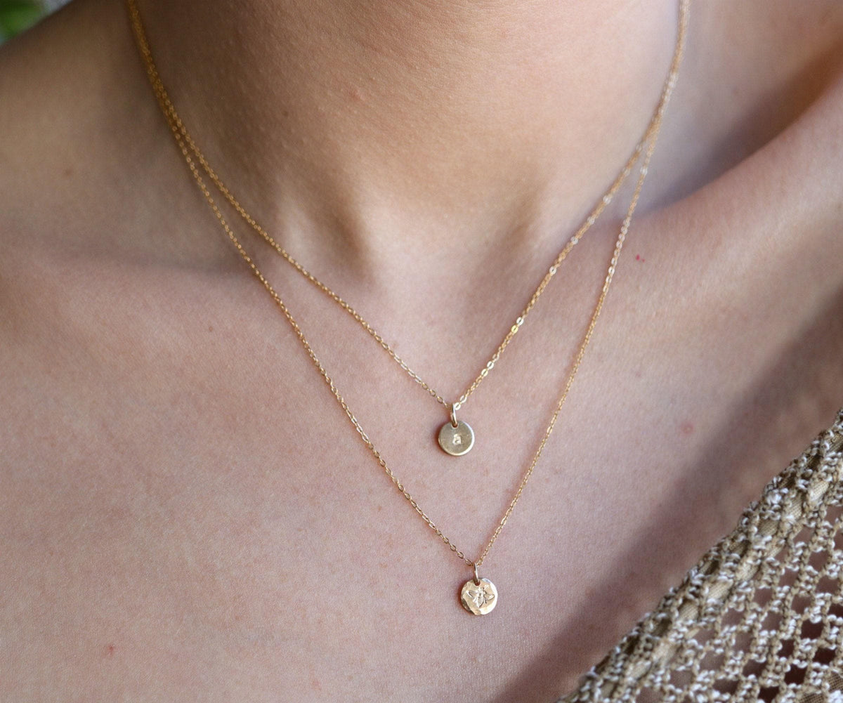 A person wearing a layered gold necklace set consisting of a short chain with an initial pendant and a longer chain with a bee pendant.