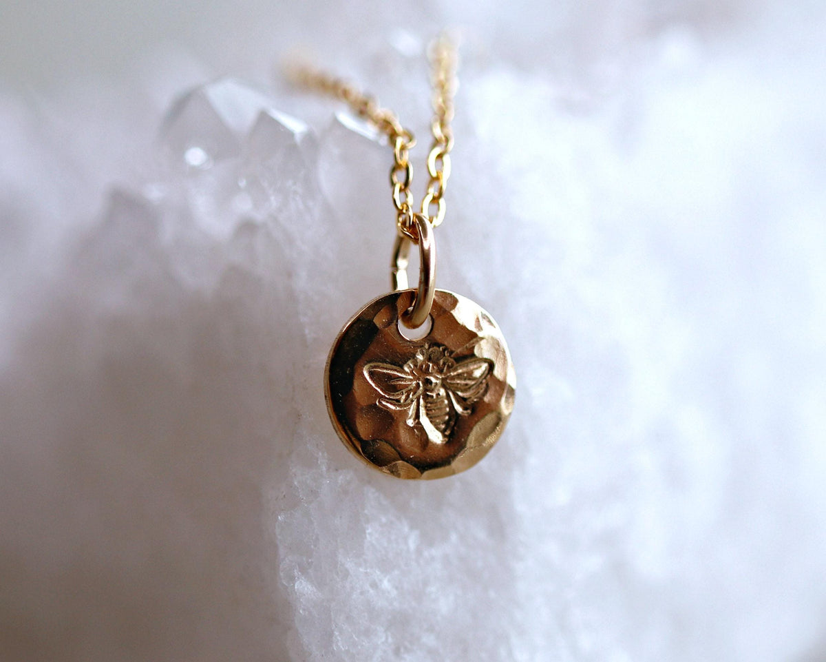 A detailed shot of a gold textured circular pendant stamped with a detailed bee design on a white crystal background.