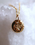 A detailed shot of a gold textured circular pendant stamped with a detailed bee design on a white crystal background.