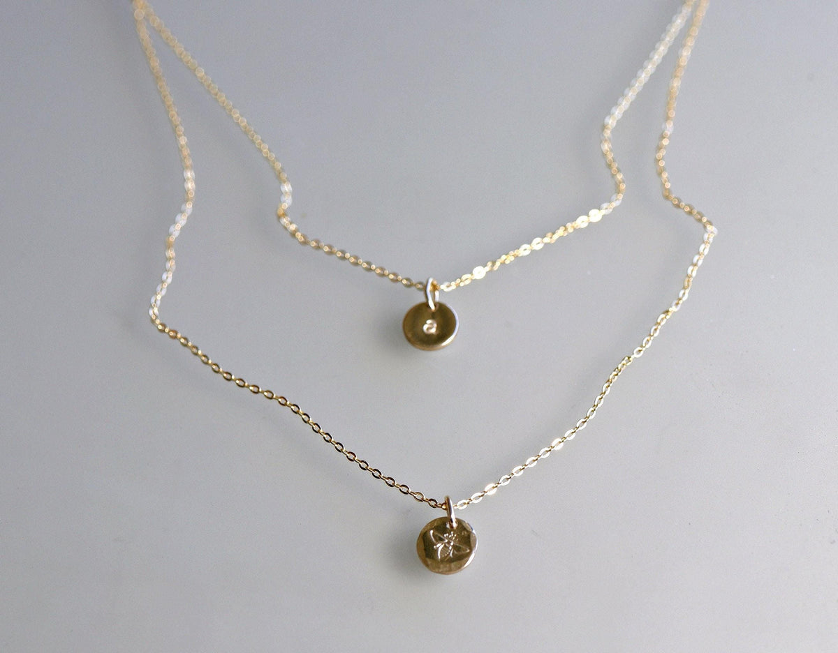 Two layered gold chain necklaces displayed flat on a neutral surface, the top necklace having a tiny initial disc and the bottom a bee disc.