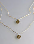 Two layered gold chain necklaces displayed flat on a neutral surface, the top necklace having a tiny initial disc and the bottom a bee disc.