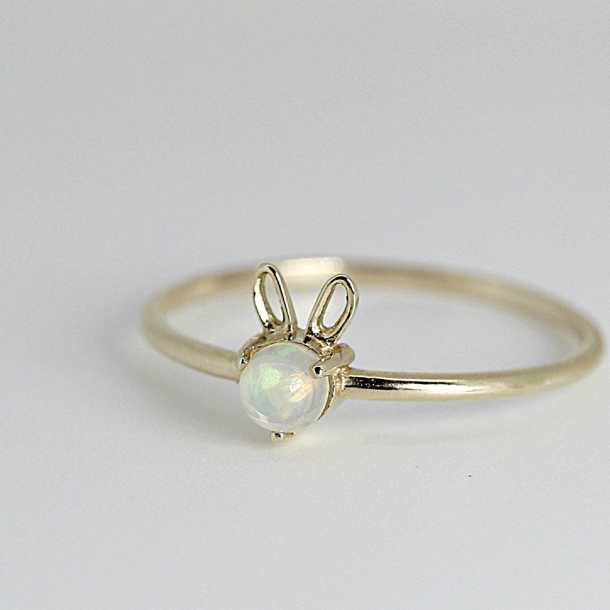 A dainty gold ring featuring a round iridescent opal and two wireform gold bunny ears.