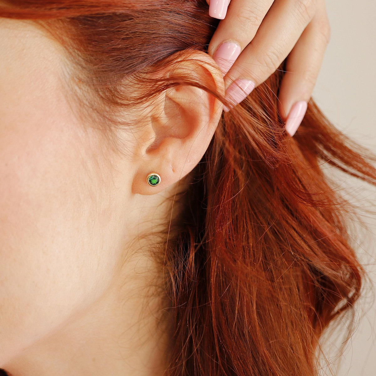 A close-up shot of a single round green emerald stud earring worn in an earlobe, with red hair framing the side of the face.