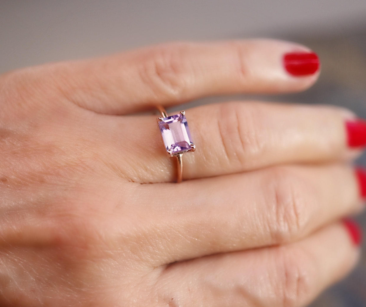 A hand with red nail polish wearing the thin gold ring with a horizontal rectangular purple gemstone.
