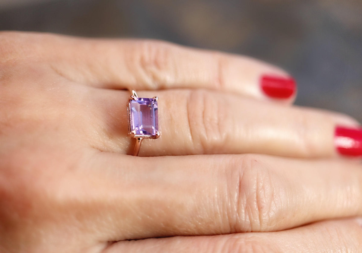 A close-up shot of a hand with red manicure wearing the thin gold ring with a vertically oriented rectangular purple gemstone.
