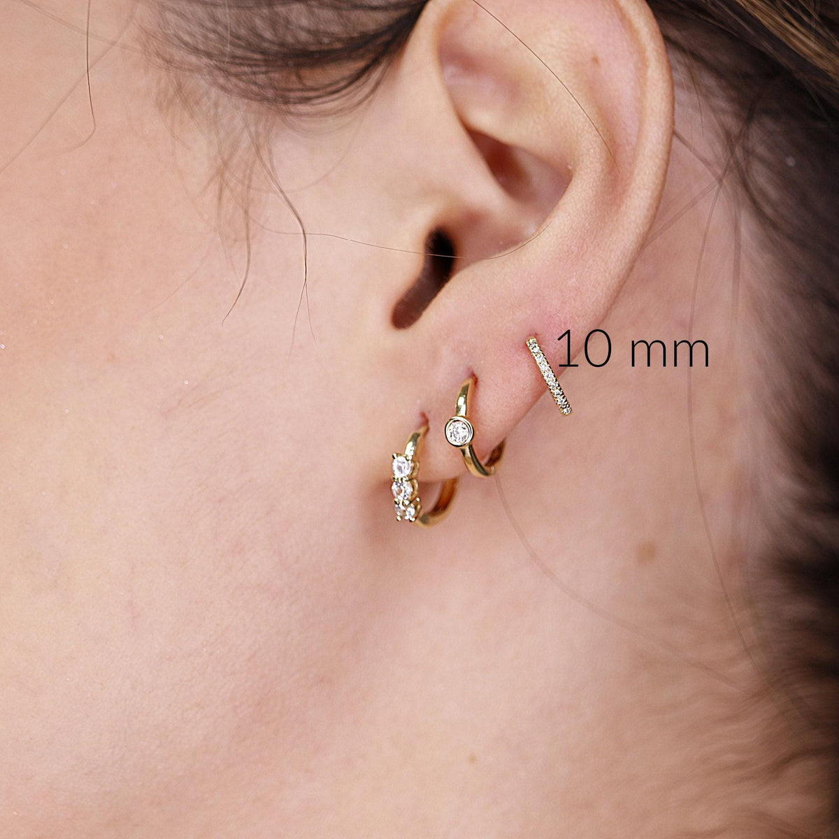 A close-up of a human ear wearing a small diamond-pave huggie hoop earring labeled 10 mm alongside two other earrings.