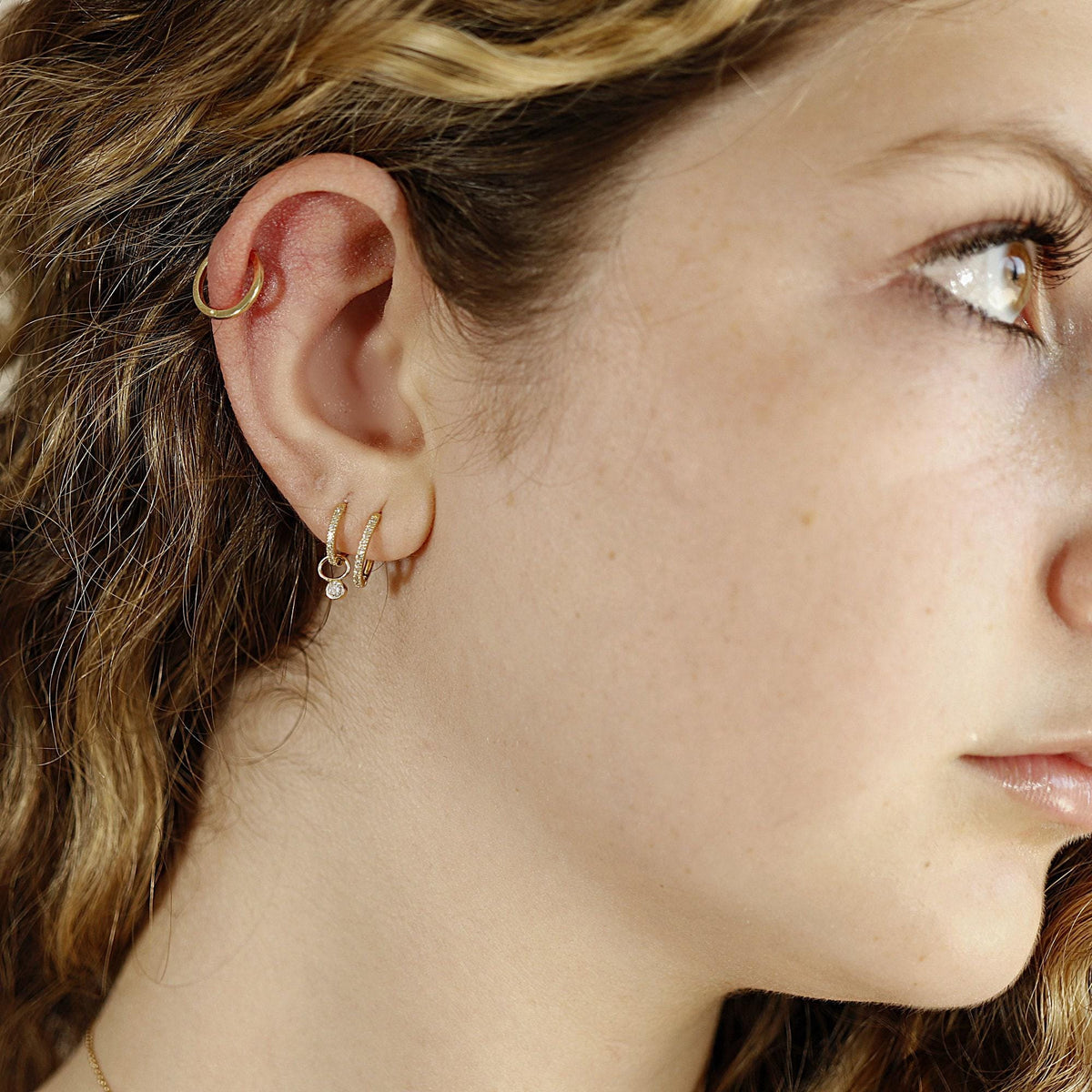 A close-up of a person wearing a plain gold hoop in the cartilage and two small pave huggie hoops with a dangling charm in the lobe.